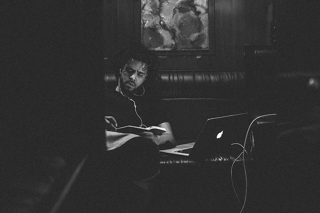 J. Cole is seen reading a book with his headphones connected. Image Owned by Michael Wolf and considered public domain work as of December 22, 2016